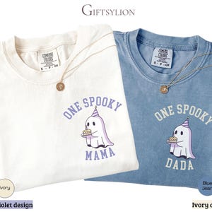May include: Two short-sleeve crew neck t-shirts, one ivory and one blue jean, each featuring a graphic of a ghost wearing a party hat and holding a birthday cake. The ivory shirt has violet text reading ONE SPOOKY MAMA, while the blue shirt has ivory text reading ONE SPOOKY DADA.