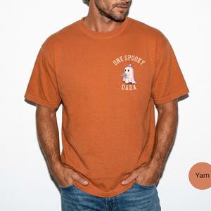 May include: A burnt orange short-sleeve t-shirt featuring a small graphic on the left chest. The graphic depicts a white ghost wearing a purple party hat and holding a small brown object, with the text ONE SPOOKY DADA printed in a curved beige font.