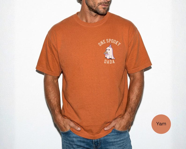 May include: A burnt orange short-sleeve t-shirt featuring a small graphic on the left chest. The graphic depicts a white ghost wearing a purple party hat and holding a small brown object, with the text ONE SPOOKY DADA printed in a curved beige font.