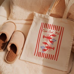 May include: A pair of beige plush slippers with brown textured insoles and a cream-colored canvas tote bag featuring a red striped design, two red lobster illustrations, blue cocktail graphics, and the text Cape Cod, MA printed in red.
