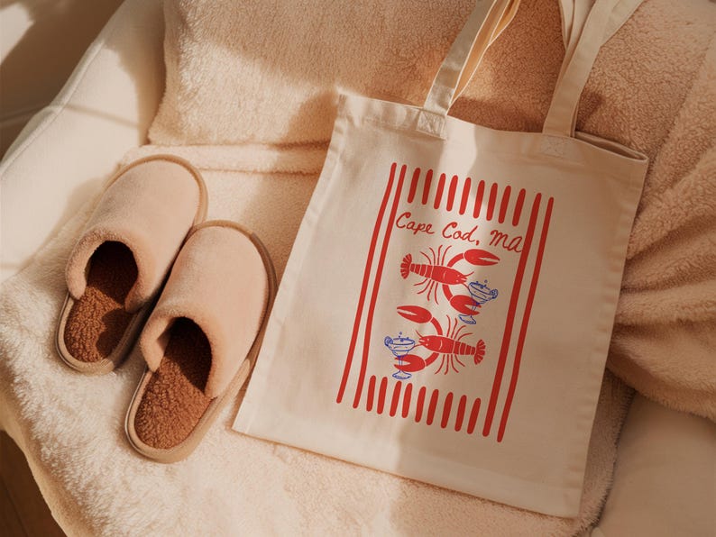 May include: A pair of beige plush slippers with brown textured insoles and a cream-colored canvas tote bag featuring a red striped design, two red lobster illustrations, blue cocktail graphics, and the text Cape Cod, MA printed in red.