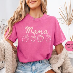 Op de afbeelding: Een felroze t-shirt met het woord "Mama" en vier cr&egrave;mekleurige aardbeienillustraties. Het shirt heeft een ronde hals en korte mouwen. Het model draagt een blauwe spijkerbroek en een cr&egrave;me jas.