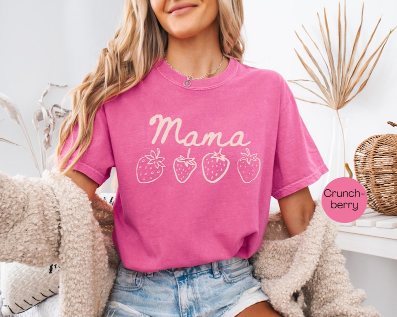 Op de afbeelding: Een felroze t-shirt met het woord "Mama" en vier cr&egrave;mekleurige aardbeienillustraties. Het shirt heeft een ronde hals en korte mouwen. Het model draagt een blauwe spijkerbroek en een cr&egrave;me jas.