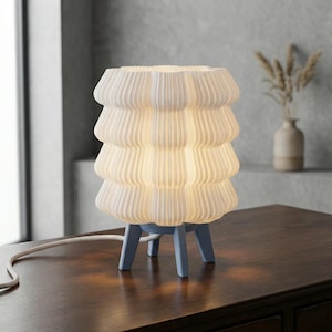 Op de afbeelding: Een witte, gestapelde, geribbelde keramische tafellamp met een zachte gloed. De lamp staat op een lichtblauwe, driepotige voet. Het snoer van de lamp is wit. De lamp staat op een donkerbruine houten tafel.