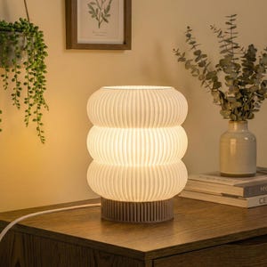 Op de afbeelding: Een witte tafellamp van geribbeld glas met een gestapeld, afgerond ontwerp. De lamp is verlicht en geeft een warme gloed. Hij staat op een houten oppervlak, met een zichtbaar netsnoer. De lamp is een decoratieve verlichtingsarmatuur.