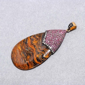 May include: A teardrop-shaped pendant with a polished, marbled surface in shades of orange, brown, and black. The top section is a triangular shape covered in small, pink gemstones, bordered by a row of white gems. The bail is gold-coloured.