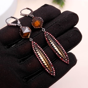 May include: A pair of dangle earrings with a dark metal finish. Each earring features a faceted, amber-colored stone at the top, connected to a long, teardrop-shaped element. The teardrop is outlined with small red stones and has a gold-colored center.