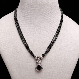 Puede incluir: Un collar de cuentas negras con un colgante plateado. El colgante presenta una piedra triangular negra rodeada de pequeñas piedras transparentes. El collar se exhibe en un maniquí blanco.