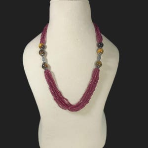 May include: A multi-strand necklace on a white mannequin. It features small, round, deep pink beads and larger, round tiger's eye and black beads. The necklace is knotted at the bottom. The beads are measured in centimetres.