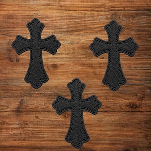 Puede incluir: Tres parches negros con forma de cruz que presentan una superficie texturizada y granulada con costuras decorativas en los bordes. Cada cruz tiene una forma ornamental con extremos ensanchados, dispuesta en una formación triangular.