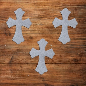 May include: Three light gray cross-shaped cutouts with a textured surface. Each cross features a decorative, ornate silhouette with rounded edges and flared ends, arranged in a triangular formation against a brown wood grain background.