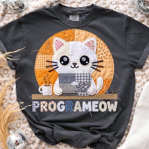 May include: Dark gray t-shirt featuring a patchwork cat design with large eyes, whiskers, and a coffee cup. The cat is set against an orange and yellow background. The text "PROGRAMEOW" is displayed below the cat.