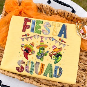 Può includere: Una maglietta gialla con le parole "FIESTA SQUAD" in lettere patchwork colorate. La maglietta presenta immagini ricamate di peperoncini e cactus con sombrero che suonano strumenti. La maglietta è esposta in un cestino di vimini con tessuto arancione.