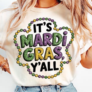 May include: A cream-coloured t-shirt featuring the text "IT'S MARDI GRAS Y'ALL" in green, purple, and yellow glitter lettering. The text is framed by a beaded necklace design in matching colours. A festive design for Mardi Gras celebrations.