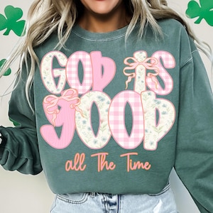 May include: A teal green sweatshirt with the phrase "GOD IS GOOD all The Time" in a mix of pink gingham, floral, and cursive text. The letters have a 3D effect with bows and floral accents.