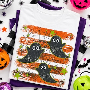 May include: A white t-shirt featuring a graphic print of three black ghosts floating over a black spiderweb design. The background of the print consists of horizontal orange and brown distressed brush strokes accented with small lime green stars.