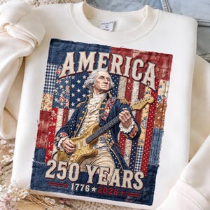 May include: Cream-colored sweatshirt featuring a graphic of George Washington playing a guitar. The graphic includes the word "AMERICA" and "250 YEARS 1776-2026" on a patchwork design of the American flag. The design is in red, white, and blue.