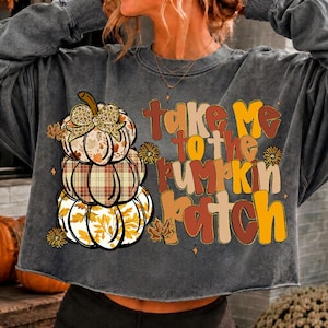 May include: A charcoal gray long-sleeve sweatshirt featuring a graphic of three stacked pumpkins with leopard print, plaid, and leaf patterns. The text "take me to the pumpkin patch" is printed in a multi-colored, stylized font alongside small leaf and flower icons.