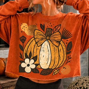 May include: A burnt orange long-sleeve sweatshirt featuring a graphic print of a large pumpkin with a bow on top, surrounded by stylized leaves and a white flower. The design includes abstract circular shapes and dark brown line details.
