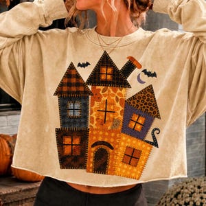 May include: A beige long-sleeve sweatshirt featuring a graphic of a multi-sectioned house with various patterns including buffalo plaid, leopard print, and polka dots. The house design includes orange windows, a chimney, and two black bats.