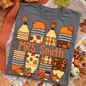 Op de afbeelding: Een donkergrijs t-shirt met een grafische print van meerdere potloden met herfstthema-patronen, waaronder oranje, bruine en groene bladeren, ruitjes en chevronontwerpen. De tekst "MRS. Smith" is in het oranje in het midden gedrukt.