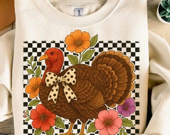 Pode incluir: Camisola creme com uma ilustração detalhada de um peru com uma gravata borboleta de bolinhas, rodeado por flores coloridas. O peru está sobre um fundo xadrez preto e branco, criando um design festivo e sazonal.
