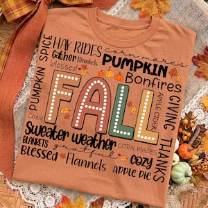 May include: A burnt orange t-shirt featuring the word FALL in large, dotted block letters surrounded by autumn-themed text including Hay Rides, Pumpkin, Bonfires, Apple Cider, Sweater weather, Corn Mazes, Blessed, Grateful, and Flannels.
