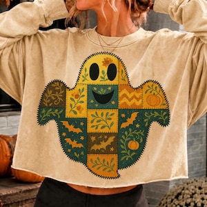 May include: A beige long-sleeve sweatshirt featuring a large, central ghost graphic composed of a patchwork quilt design. The ghost pattern includes various autumn-themed motifs such as pumpkins, bats, and leafy vines in shades of orange, green, and brown.