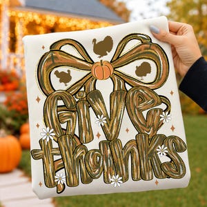 May include: A white sweatshirt featuring a large graphic with the text Give Thanks in a textured brown and green pattern. The design includes a prominent bow, a central orange pumpkin, three brown turkey silhouettes, and small white daisies.
