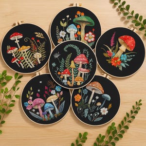 Puede incluir: Seis obras de arte de hongos bordados en aros de madera. Cada una presenta diseños de hongos coloridos con acentos florales y de follaje sobre un fondo negro. El bordado incluye detalles rojos, naranjas, azules y blancos, creando un tema caprichoso inspirado en la naturaleza.