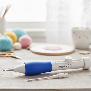 Puede incluir: Una herramienta de aguja de perforación blanca y azul con un agarre azul y una punta de aguja. La herramienta tiene una escala numerada y está junto a una aguja más pequeña. El fondo incluye hilo, un bastidor de bordado y una taza de té.