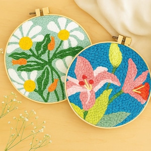 May include: Two circular embroidery hoops with floral designs. One shows white daisies with yellow centres and green leaves on a light blue background. The other has pink lilies on a blue background with green leaves.