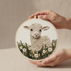 Op de afbeelding: Geborduurd kunstwerk van een lammetje in een veld met madeliefjes, in een houten borduurring. Het lam is beige met een getextureerd uiterlijk, omgeven door groene bladeren en witte en gele bloemen. Het kunstwerk wordt door handen vastgehouden.