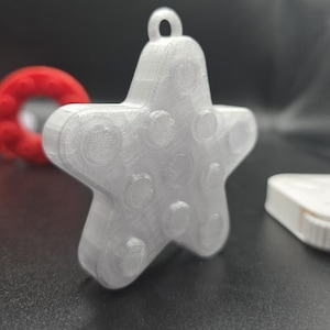 May include: A white, star-shaped ornament with a loop for hanging. The star has a textured surface with circular indentations. A red ring and a white object are partially visible in the background.