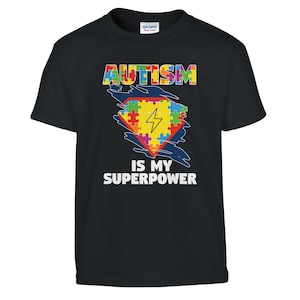 May include: A black short-sleeved t-shirt featuring a graphic of a shield filled with a colourful puzzle piece pattern and a yellow lightning bolt. The text reads AUTISM IS MY SUPERPOWER in bold, colourful, and white lettering on the front.