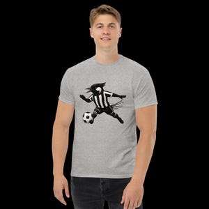 May include: A light gray t-shirt featuring a cartoon magpie playing soccer. The magpie is wearing a black and white striped uniform and kicking a soccer ball. The shirt is a classic crew neck style.