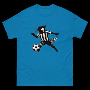 May include: A turquoise blue t-shirt featuring a cartoon bird in a soccer uniform, dribbling a soccer ball. The bird is black and white, wearing a striped jersey and shorts. The soccer ball is black and white.