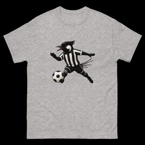 May include: A gray t-shirt featuring a cartoon bird in a black and white soccer uniform, kicking a soccer ball. The bird has a black head and beak, with a white face. The shirt is made of a soft, comfortable material.