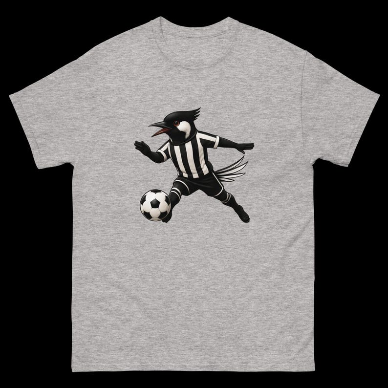 May include: A gray t-shirt featuring a cartoon bird in a black and white soccer uniform, kicking a soccer ball. The bird has a black head and beak, with a white face. The shirt is made of a soft, comfortable material.