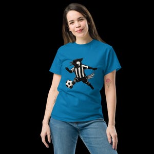 May include: A turquoise t-shirt featuring a cartoon magpie playing soccer. The magpie is dressed in a black and white striped uniform and is kicking a soccer ball. The shirt is made of a soft material.