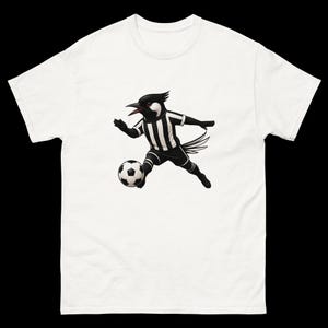 May include: White t-shirt featuring a cartoon bird in a black and white soccer uniform, dribbling a soccer ball. The bird has a black head and beak, with a striped jersey and shorts. The soccer ball is black and white.