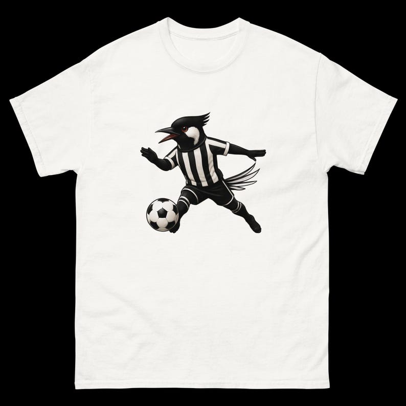 May include: White t-shirt featuring a cartoon bird in a black and white soccer uniform, dribbling a soccer ball. The bird has a black head and beak, with a striped jersey and shorts. The soccer ball is black and white.
