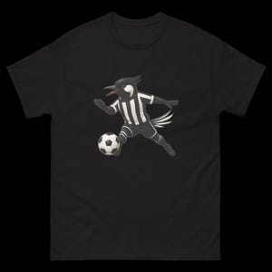 May include: Black t-shirt featuring a cartoon magpie in a soccer uniform, kicking a soccer ball. The magpie is black and white, with a striped jersey and shorts. The soccer ball is black and white.