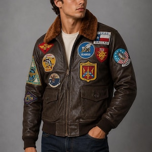 May include: A brown leather bomber jacket featuring a brown faux fur collar, a front zipper, and multiple colorful embroidered military-style patches on the chest and sleeves, including patches labeled Texas, Seabees, and USS William H. Standley.