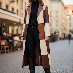 Puede incluir: Un abrigo largo con bloques de color marrón, crema y marrón oscuro. El abrigo tiene solapas clásicas y cierre de botones. La persona lleva un cuello alto negro, vaqueros negros y botas altas negras. El fondo es una calle europea.