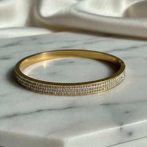 May include: Gold-toned bangle bracelet with three rows of small, sparkling crystals. The bracelet is set against a marble surface with a soft, white fabric in the background. A simple, elegant piece of jewelry.