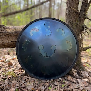 May include: A handpan drum with a dark blue and black finish. The steel drum has several circular cutouts and is surrounded by a black rubber ring. The instrument is resting on a bed of leaves and twigs in a forest setting.