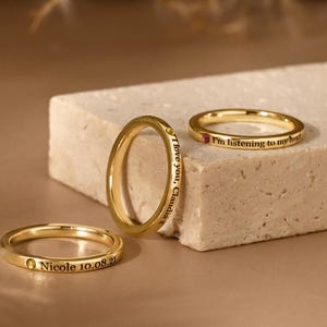 Op de afbeelding: Drie goudkleurige ringen worden getoond op een beige oppervlak. Elke ring heeft een dunne band en gegraveerde tekst. Eén ring leest "Nicole 10.08.21", een andere zegt "I love you, Claudia", en de derde zegt "I'm listening to my heart."