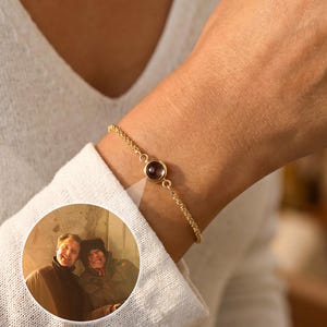 Può includere: Un braccialetto color oro con una piccola gemma rotonda di colore rosso scuro. Il braccialetto è su un polso e una piccola immagine circolare di due persone è visibile nelle vicinanze. Il braccialetto ha un delicato design a catena.