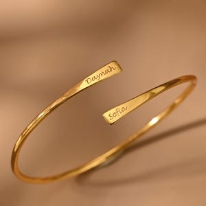 Peut inclure: Un bracelet manchette ouvert de couleur or, avec les noms "Daynah" et "Sofia" gravés aux extrémités. Le bracelet a une texture martelée et est présenté sur une surface aux tons neutres.